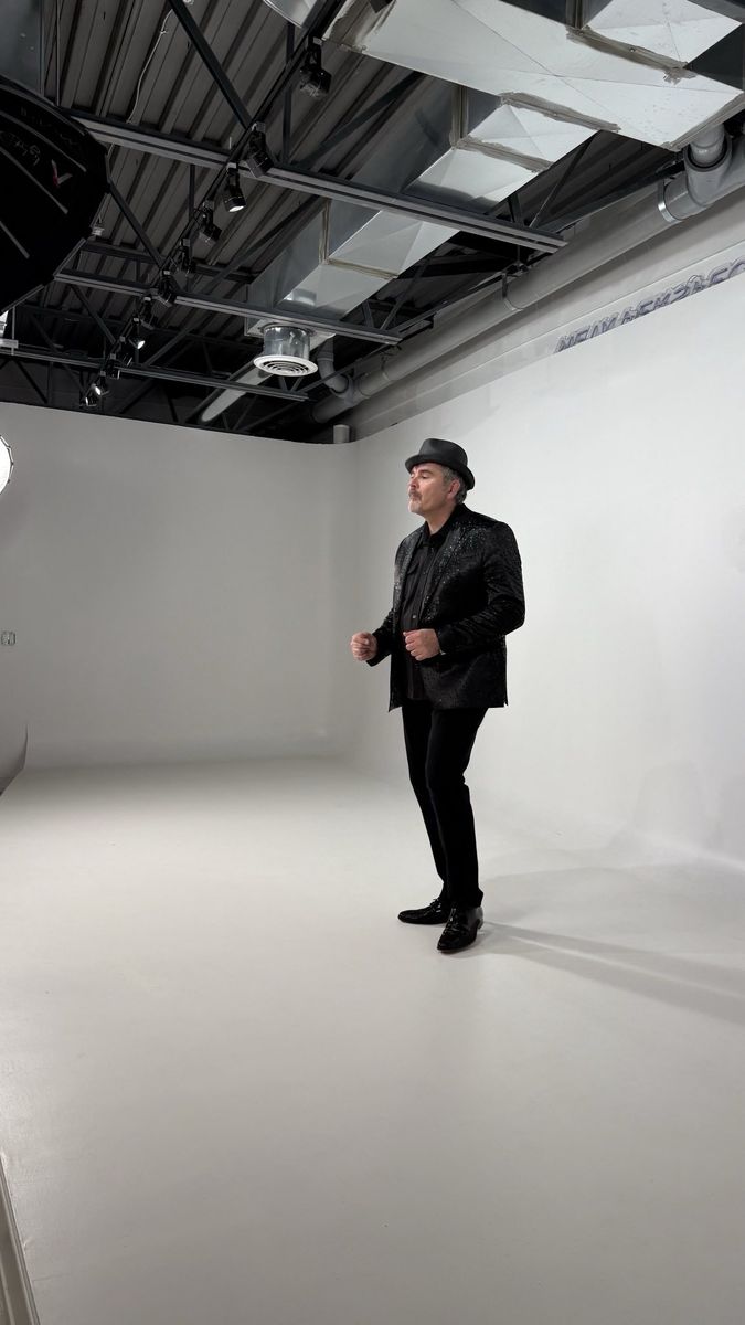 Tristan in a black sparkling jacket and fedora standing in a bright white studio