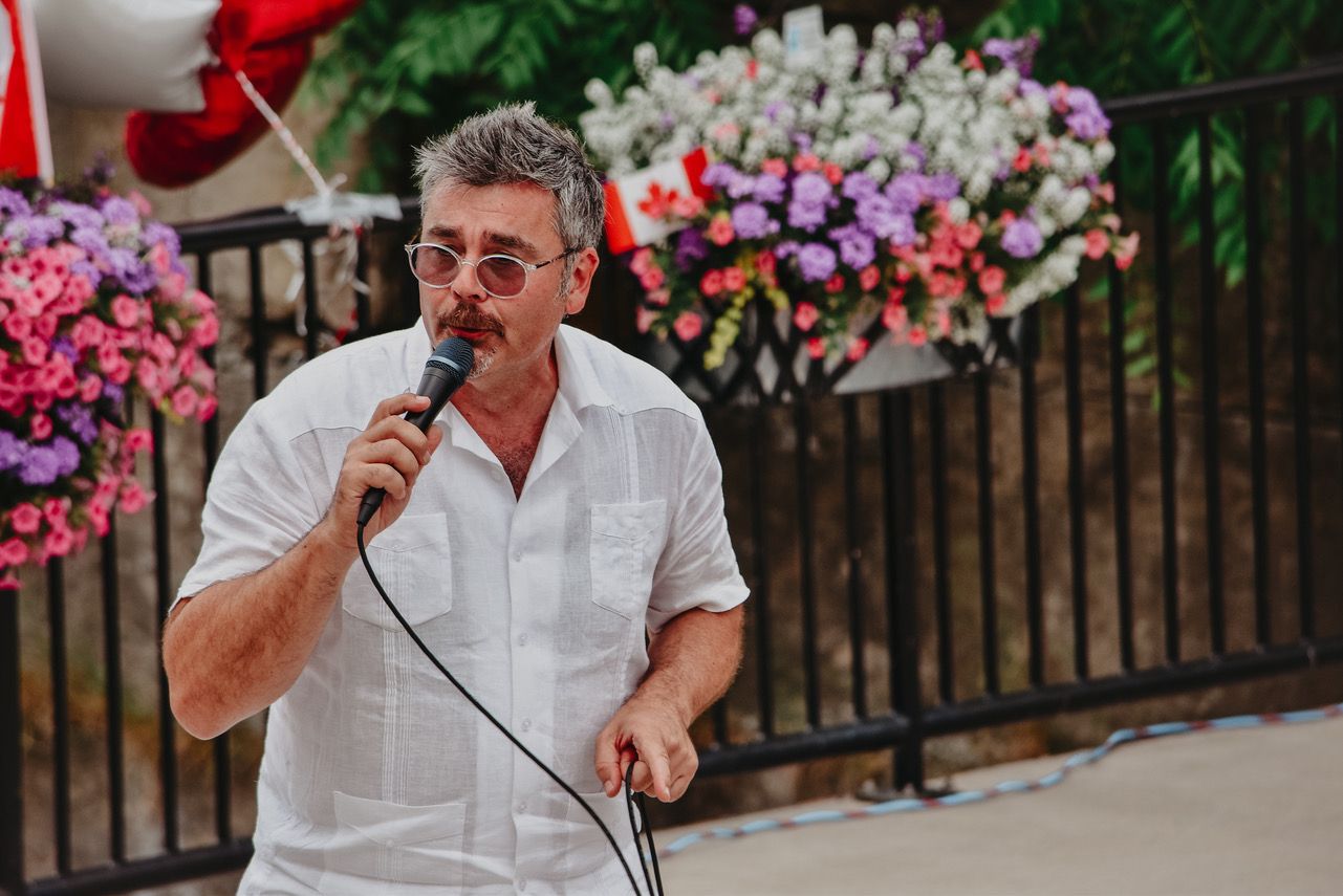 Tristan in a white shirt singing into a microphone at an outdoor show, with hanging flower baskets and a Canadian flag behind him