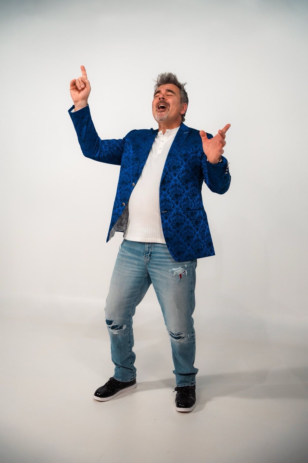 Tristan Alexander singing joyfully with one finger pointed skyward, wearing a blue paisley blazer, white T-shirt and jeans